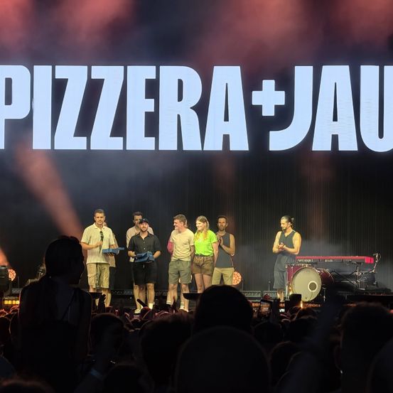 Eine Bühnenaufführung mit sechs Personen, die vor einem Publikum stehen, Pokale halten und lächeln. Das Wort 'PIZZERA+JAU' ist prominent auf der Bühne zu sehen.