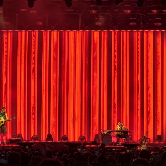 Zwei Musiker spielen auf einer Bühne mit rotem Vorhang und Publikum vor ihnen.