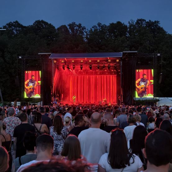 Ein großes Publikum sieht eine Live-Aufführung auf einer Bühne mit roter Beleuchtung und großen Bildschirmen, die den Künstler zeigen. Die Bühne ist vor einem Hintergrund von Bäumen aufgebaut.