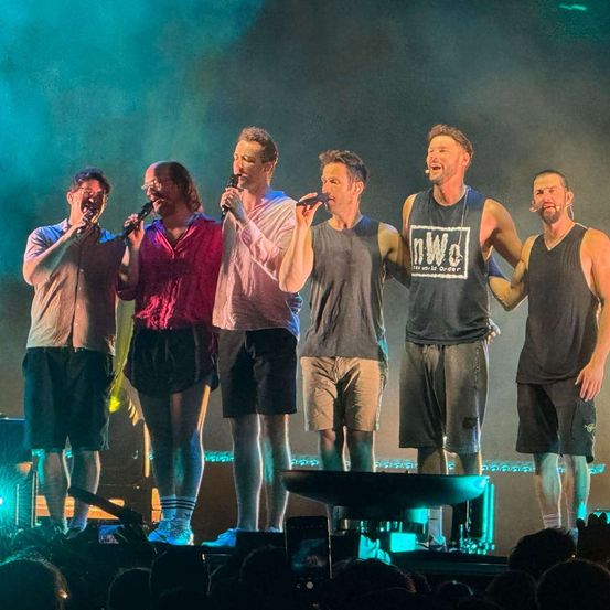 Sechs Männer stehen mit Mikrofonen singend auf der Bühne. Sie tragen lässige Kleidung, drei tragen ärmellose Shirts und die anderen drei tragen Hemden. Dahinter steht eine große Menge.