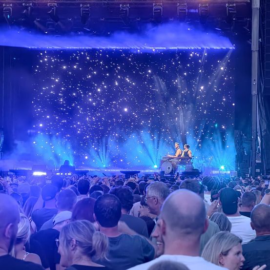 Ein Publikum sieht eine Live-Aufführung auf der Bühne bei blauer Beleuchtung mit Sternenhimmel als Hintergrund. Zwei Künstler spielen Instrumente.