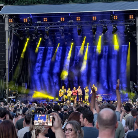 Eine Band spielt auf einer Bühne mit hellen gelben und blauen Lichtern. Das Publikum vor der Bühne macht Fotos und jubelt.