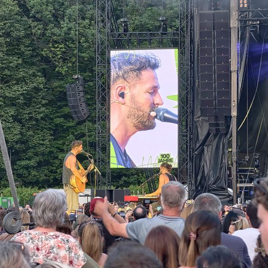 Ein Musiker spielt Gitarre auf der Bühne mit einer großen Leinwand im Hintergrund, die sein Bild zeigt. Ein Publikum beobachtet von vorne.