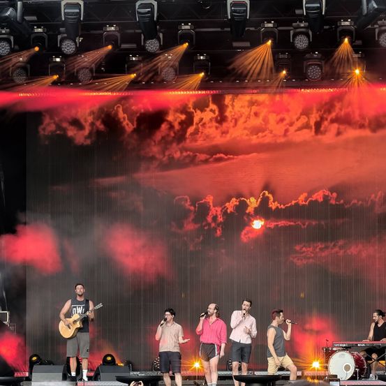 Eine Band tritt auf der Bühne auf, mit dramatischer roter Beleuchtung und Raucheffekten. Ein Mann spielt Gitarre, während andere singen und Instrumente spielen.