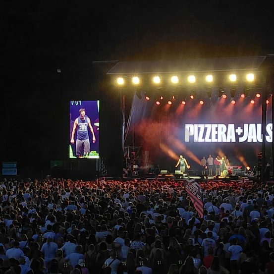 Ein nächtliches Ereignis zeigt einen Mann auf der Bühne mit 'PIZZERIA+JALS' beleuchtet. Viele Menschen schauen zu, und die Bühne ist mit roten Lichtern beleuchtet.