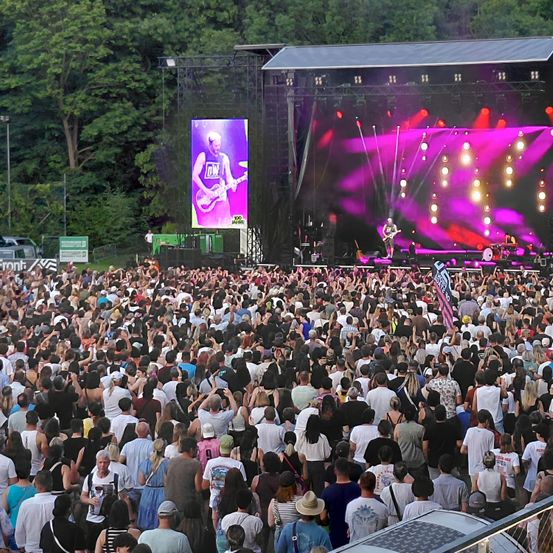 Eine große Menschenmenge versammelte sich bei einem Open-Air-Musikfestival. Die Bühne ist mit roten und lila Lichtern beleuchtet. Ein Musiker spielt Gitarre auf der Bühne.