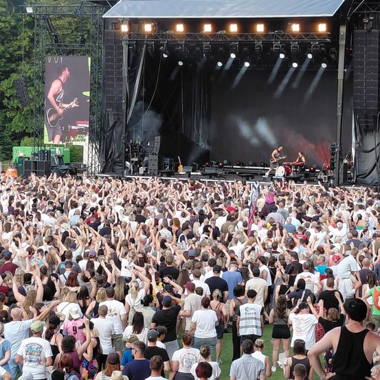 Eine große Menschenmenge versammelt sich, um ein Konzert auf der Bühne zu sehen, wobei ein Gitarrist auf einer großen Leinwand im Hintergrund spielt. Raucheffekte und Beleuchtung erhöhen die Atmosphäre.