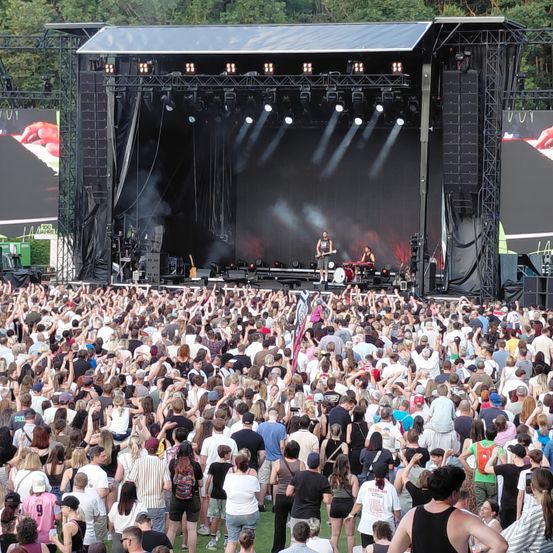 Ein großes Open-Air-Musikfestival mit einer Bühne, auf der zwei Künstler auftreten, einer spielt Gitarre und der andere sitzt mit einem Schlagzeug, umgeben von Nebel. Das Publikum ist dicht gedrängt und erhebt die Hände, um die Show zu genießen.
