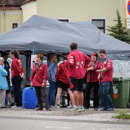 Eine Gruppe von Menschen in roten Jacken versammelt sich unter einem Zelt, einige mit Abzeichen und einige im Gespräch. Ein blauer Kanister und ein grüner Mülleimer sind in der Nähe.