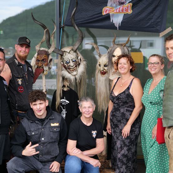 Gruppenfoto von Menschen mit Masken und Ziegenköpfen. Ein Banner mit dem Wort 'Teufel' ist im Hintergrund.
