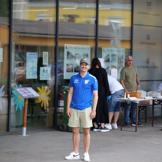 Ein Mann in einem blauen Shirt mit Baseballkappe lächelt vor einer Glaswand mit Werbung. Einige Leute stehen hinter ihm. Es gibt einen Tisch mit Essen und einem Korb.