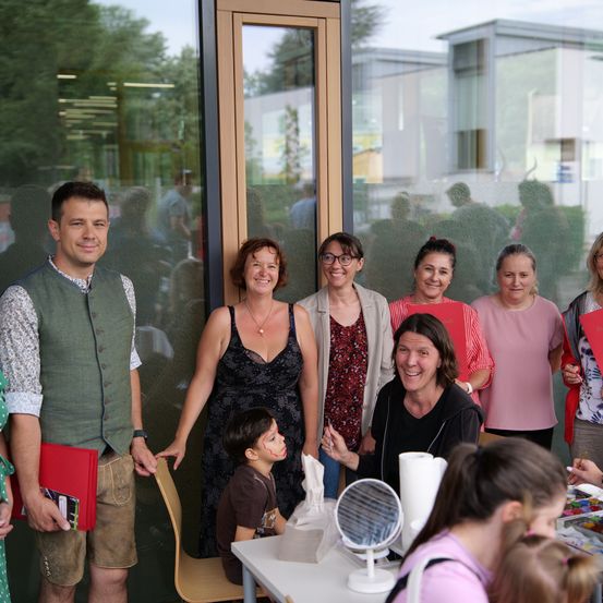 Eine Gruppe von Erwachsenen und Kindern steht gemeinsam vor einem Gebäude und lächelt für ein Foto. Ein Kind sitzt an einem Tisch mit Malutensilien.