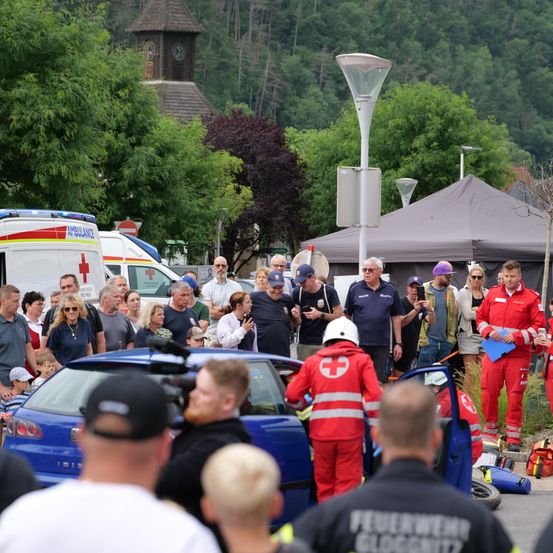 Rettungskräfte und Schaulustige versammeln sich um die Unfallstelle eines blauen Autos. Feuerwehrleute in roten Uniformen und ein Mann in einem weißen Helm kümmern sich um das Fahrzeug.