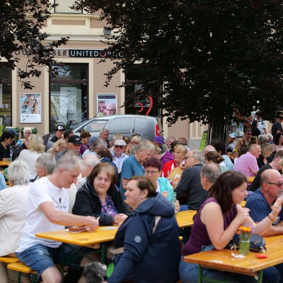 Eine Menschenmenge sitzt um Tische herum draußen vor einem Gebäude. Einige essen, während andere sich unterhalten. Ein Van ist in der Nähe geparkt.