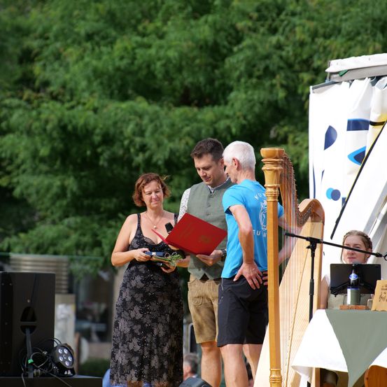 Drei Personen stehen auf der Bühne vor Bäumen und einem Mikrofon. Eine Frau hält ein rotes Buch und einen Blumenstrauß. Ein Mann spielt eine Harfe. Eine Frau sitzt an einem Tisch.
