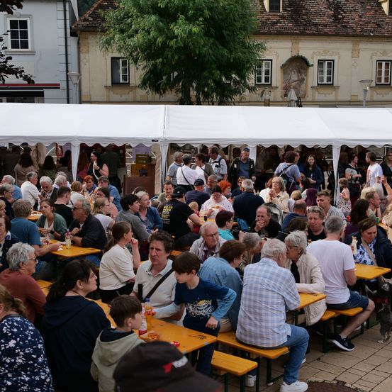 Eine Menschenmenge versammelt sich im Freien unter weißen Zelten. Viele sitzen an Tischen, essen und unterhalten sich. Das Gebiet ist von Gebäuden umgeben, eines davon mit einem Wandbild.