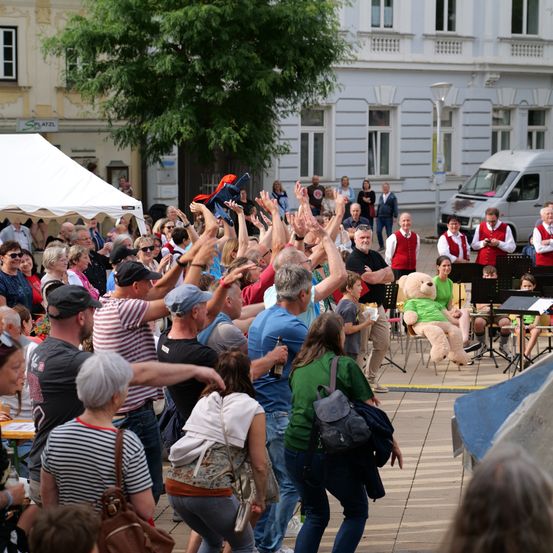 Eine Menschenmenge versammelt sich auf einer offenen Fläche vor Gebäuden. Eine Person hält einen Teddybären in die Höhe, während andere klatschen. Eine Band spielt im Hintergrund.