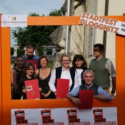 Eine Gruppe von Menschen posiert für ein Foto beim Stadtfest Gloggnitz, mit Urkunden in der Hand, vor einer Statue.