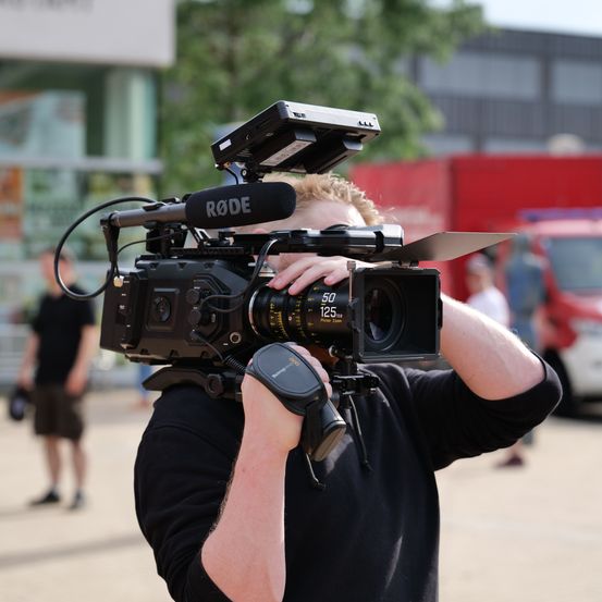 Ein Mann filmt mit einer Kamera in einem urbanen Umfeld. Er hält die Kamera mit einem Rode-Mikrofon. Im Hintergrund befinden sich Gebäude und Menschen.
