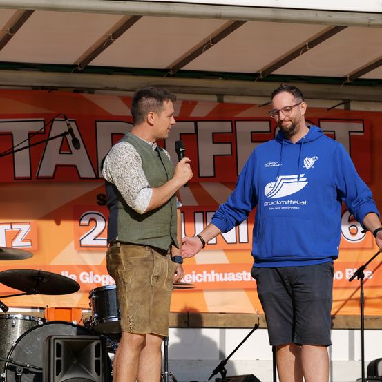 Zwei Männer auf der Bühne mit einem Mikrofon. Einer trägt eine braune Weste und Shorts, der andere eine blaue Kapuzenjacke. Dahinter steht ein Schlagzeug und ein orangefarbenes Banner.