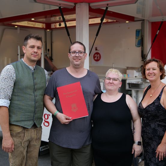 Drei Erwachsene stehen lächelnd vor einem Imbisswagen. Ein Mann in einem Weste hält ein rotes Buch. Die Frau neben ihm trägt eine Brille und ein schwarzes Tanktop. Eine weitere Frau trägt ein Kleid und eine Halskette.