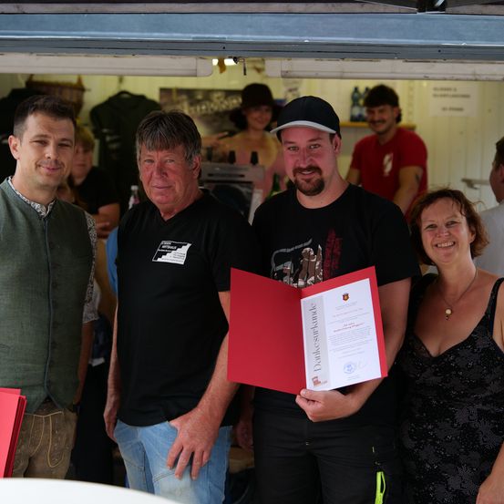 Vier Personen stehen vor einem Stand mit einer roten Mappe in der Hand des mittleren Mannes. Drei Männer und eine Frau, lächelnd. Im Hintergrund steht eine weitere Person in der Nähe eines Regals mit Flaschen.