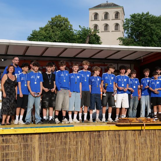Eine Gruppe junger Menschen in blauen Hemden und mit Medaillen steht auf der Bühne, neben einer Frau in Schwarz und einem Mann mit Brille. Dahinter steht ein Gebäude mit einem Turm.