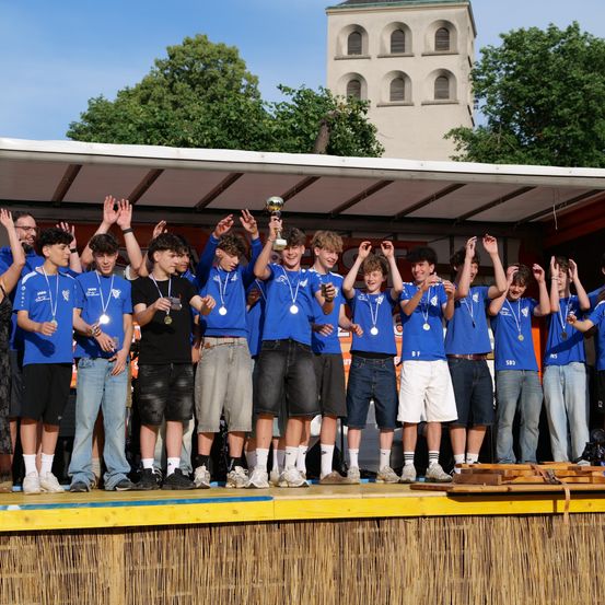 Eine Gruppe junger Männer in blauen T-Shirts mit Medaillen um den Hals steht auf der Bühne. Eine Person hält eine Trophäe in der Mitte. Sie heben die Hände, vielleicht feiern sie. Dahinter steht ein Gebäude mit einem Turm.