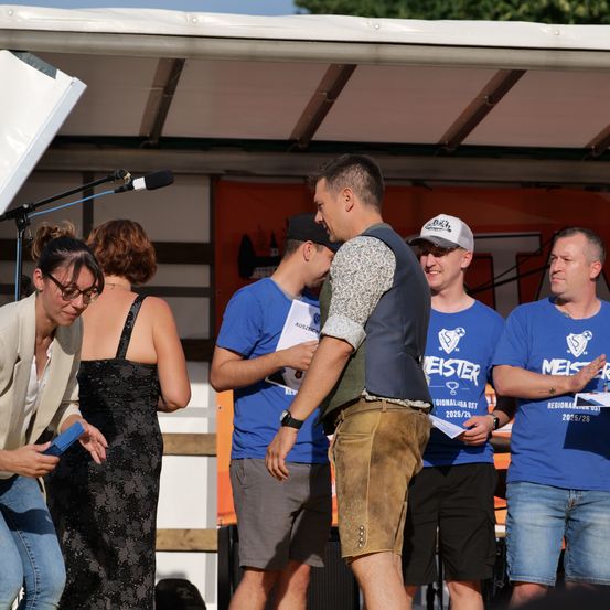 Fünf Personen stehen auf einer Bühne, zwei Männer und drei Frauen. Ein Mann im weißen Shirt hält ein blaues Objekt, möglicherweise einen Pokal. Hinter ihnen wird ein Banner angezeigt.
