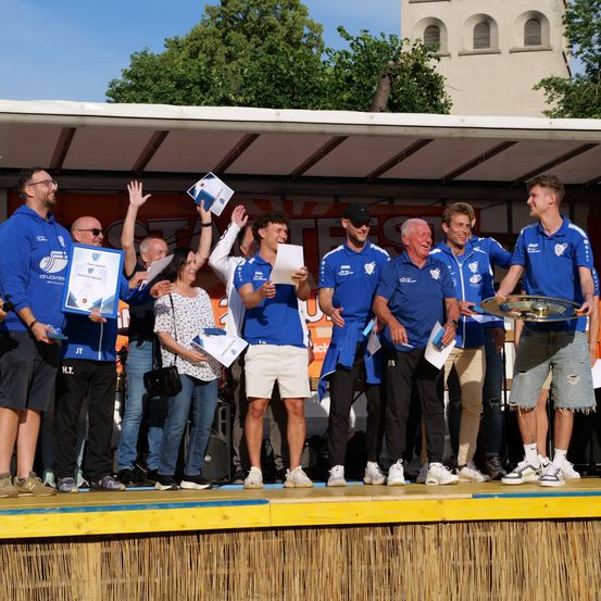 Eine Gruppe von Menschen in blauen Uniformen und mit Urkunden in den Händen steht auf einer Bühne, einige mit erhobenen Händen, lächelnd und posieren für ein Foto.
