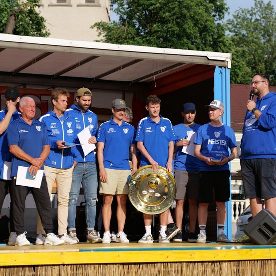 Eine Gruppe von Männern in blauen Trikots steht mit einem goldenen Pokal auf der Bühne, hält Papiere in der Hand und trägt Sneaker. Ein Mann mit einem Mikrofon spricht hinter ihnen. Bäume und ein Gebäude sind im Hintergrund.