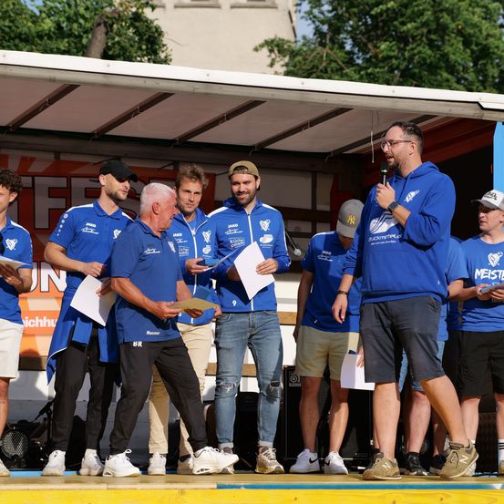 Eine Gruppe von Männern, einige mit Hüten, stehen auf einer Bühne und erhalten Auszeichnungen. Ein Mann hält ein Mikrofon. Sie tragen blaue Hemden, Hosen und Sneakers, einer trägt eine Brille. Hinter ihnen steht eine Kulisse mit Text und Bäumen.