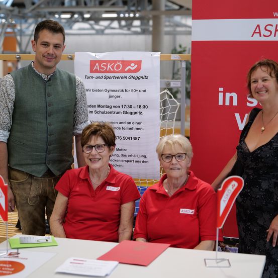 Ein Mann und zwei Frauen in roten Shirts stehen vor einem Tisch, neben einem roten Schild mit weissem Text. Der Mann trägt ein grünes Weste. Die Frauen tragen Brillen und lächeln.