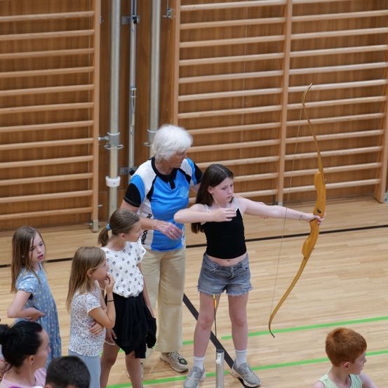 Eine Frau mit weißen Haaren zeigt einer jungen Frau, wie man einen Bogen und Pfeil benutzt, in einer Turnhalle. Andere Mädchen beobachten.