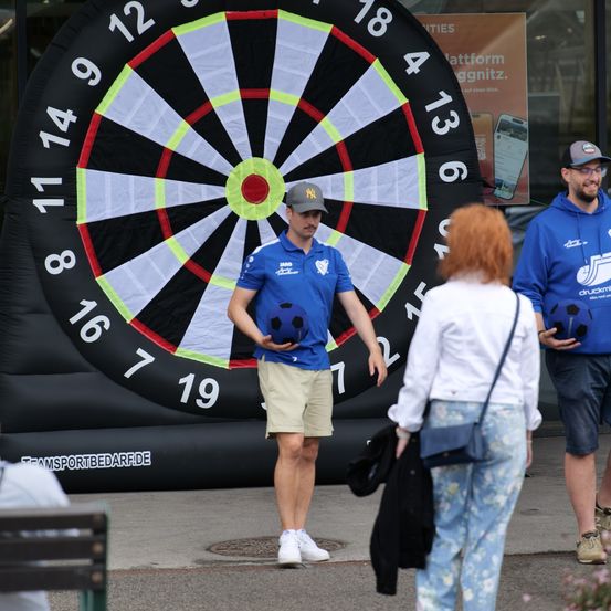 Ein Mann hält einen Fußball in der Hand und steht vor einem riesigen Dartboard. In der Nähe beobachten eine Frau in einem Blumenshirt und ein Mann in einer blauen Kapuzenjacke.