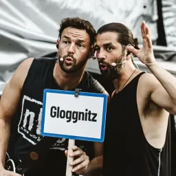 Zwei Männer in schwarzen ärmellosen Shirts und mit verkabelten Mikrofonen halten ein Schild mit der Aufschrift 'Gloggnitz'. Sie scheinen auf einer Bühne zu stehen, mit einem Publikum im Hintergrund.