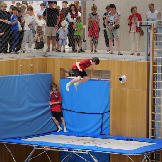 Ein Junge springt auf einem Trampolin, während andere von einer Galerie zusehen. Das Trampolin ist blau und weiß.