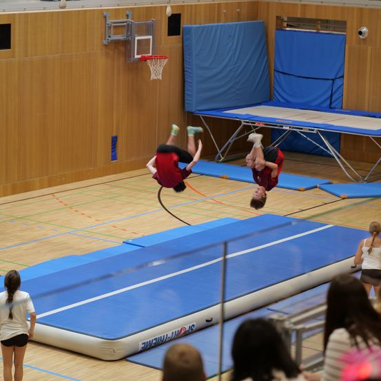 Zwei Turner führen Saltos auf einer Trampoline in einer Turnhalle aus. Einer ist in der Luft, während der andere landet. Im Hintergrund befinden sich zwei weitere Trampoline.