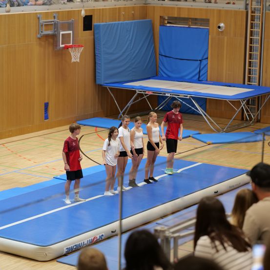Eine Gruppe von Menschen, einschließlich eines Jungen, steht auf einem blauen aufblasbaren Teppich in einer Turnhalle. Andere aufblasbare Teppiche und ein Basketballkorb sind in der Nähe. Einige Leute beobachten sie von den Seiten.