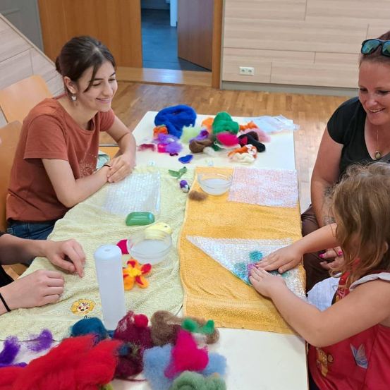 Eine Gruppe von Frauen und einem Kind sitzt um einen Tisch herum, lächelt und arbeitet an einem Bastelprojekt mit bunter Wolle und Stoff.
