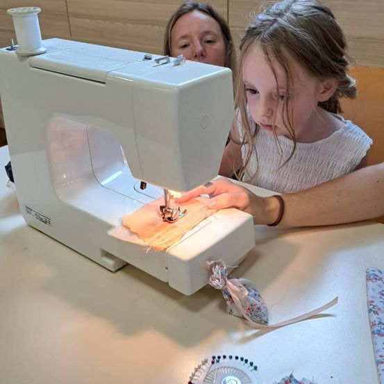 Ein junges Mädchen benutzt eine Nähmaschine mit der Hilfe einer erwachsenen Frau. Das Mädchen konzentriert sich auf ihre Aufgabe, während die Frau genau zusieht.