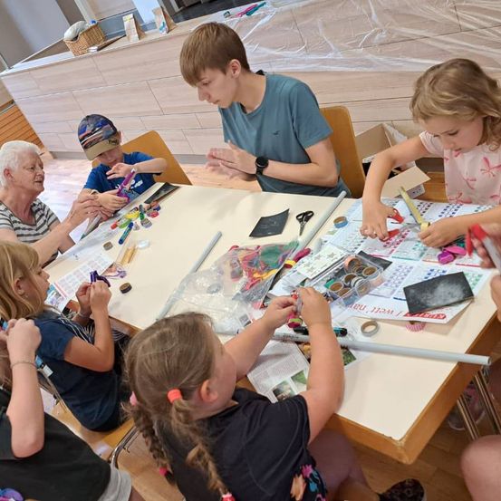 Eine Gruppe von Kindern und einem Erwachsenen versammelt sich um einen Tisch und beschäftigt sich mit einer Bastelaktivität. Sie verwenden Scheren, Kleber und andere Materialien. Der Erwachsene hilft den Kindern.