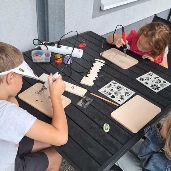 Zwei Kinder sitzen an einem Tisch, eines benutzt ein Löteisen auf einem Holzbrett, während das andere sich auf eine ähnliche Aufgabe konzentriert. Verschiedene Werkzeuge und Materialien sind auf dem Tisch verstreut.