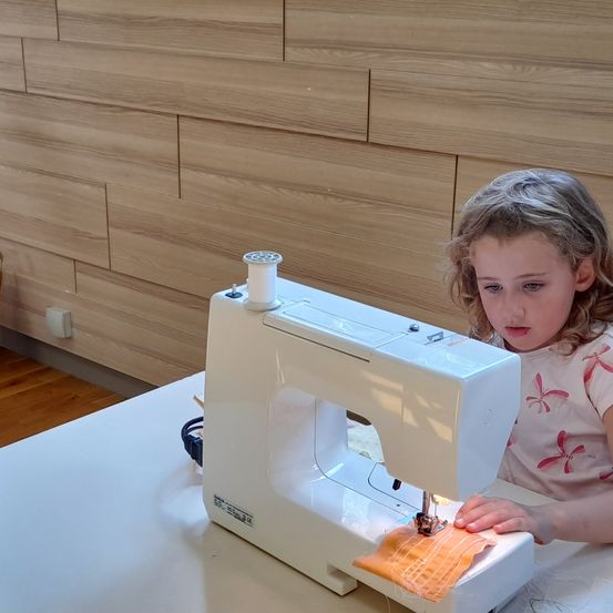 Ein kleines Mädchen sitzt an einem Tisch und benutzt eine weiße Nähmaschine, um ein orangefarbenes Stück Stoff zu nähen.