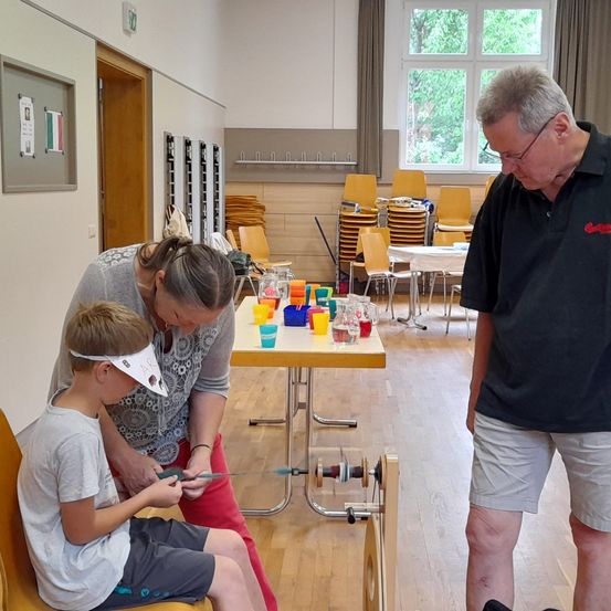 Ein junger Junge sitzt auf einem Stuhl und hält ein Werkzeug, während eine Frau hinter ihm steht und hilft. Ein Mann steht an der Seite und beobachtet. Der Raum ist mit Stühlen, einem Tisch mit verschiedenen Gegenständen und einem Fenster mit Bäumen draußen gefüllt.