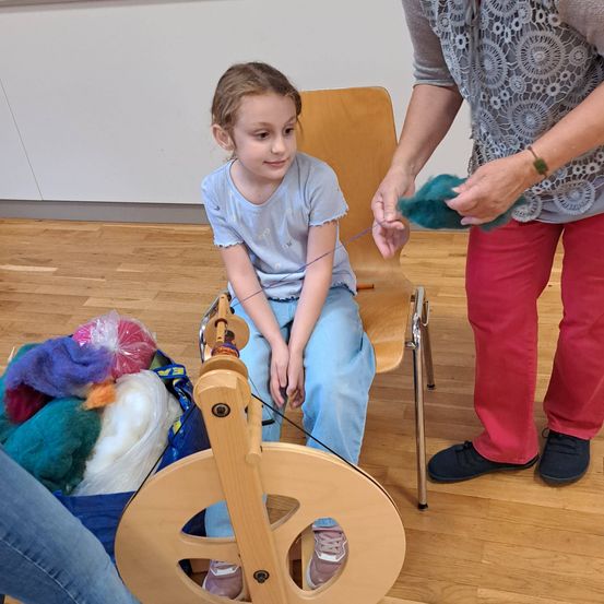 Ein kleines Mädchen in einem blauen T-Shirt und Hosen sitzt auf einem Stuhl und hält eine Spindel mit grünem Garn, während ein Erwachsener in roten Hosen sie anleitet.