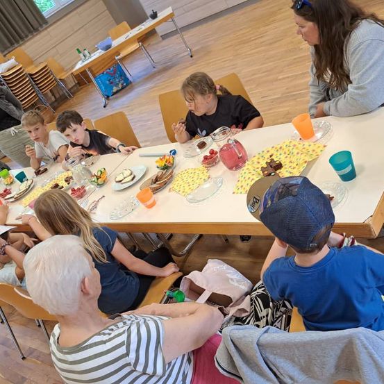 Eine Gruppe von Kindern und Erwachsenen sitzt um einen langen Tisch und genießt eine Mahlzeit. Der Tisch ist mit Tellern, Getränken und Servietten gedeckt. Die Kinder sind mit dem Essen beschäftigt, während Erwachsene beobachten. Der Raum hat Holzböden und Stühle.