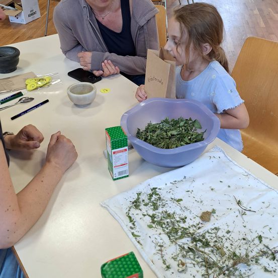 Eine Frau und ein junges Mädchen sitzen an einem Tisch, wobei das Mädchen einen Papierbeutel mit der Aufschrift 'Krautersale' hält. Sie sind von verschiedenen Gegenständen umgeben, darunter eine Schüssel mit Kräutern, eine Schachtel und ein Tuch.
