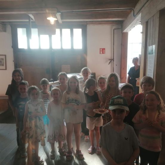 Eine Gruppe von Kindern und einigen Erwachsenen steht in einem Raum mit Holzwänden und Fenstern. Ein Kind hält einen Teller mit Essen.