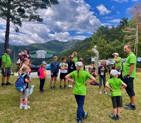 Eine Gruppe von Kindern und Erwachsenen in grünen Shirts versammelt sich auf einer Rasenfläche mit Bergen im Hintergrund. Einige tragen Hüte und haben Rucksäcke dabei.